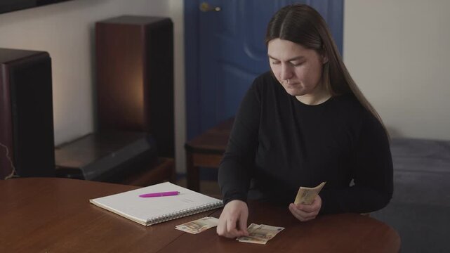 Young woman holds cash and looks uncertain while budgeting at a table, ideal for personal finance, saving money, and spending decision concepts.