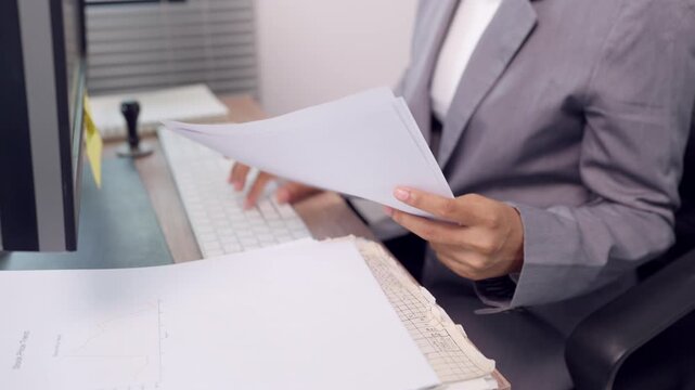 Businesswoman in formal suit reading business documents at office desk beside computer keyboard. Concept of document review, paperwork processing, office administration, and corporate workflow.