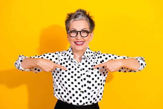 Confident mature businesswoman short grey hair glasses points to herself yellow background joy and style in a polka dot blouse for advertising and lifestyle