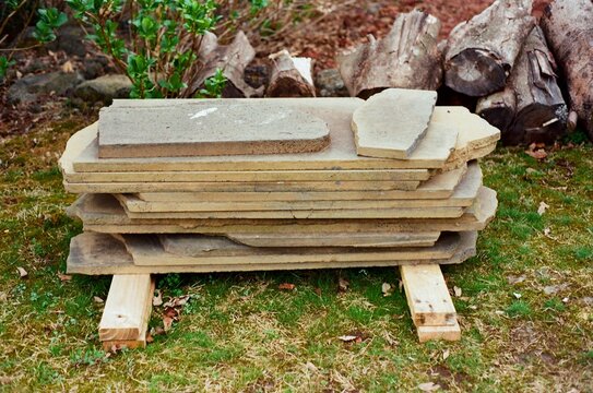 A pile of flat stone slabs stacked on wooden supports in a yard, with logs and plants in the background.