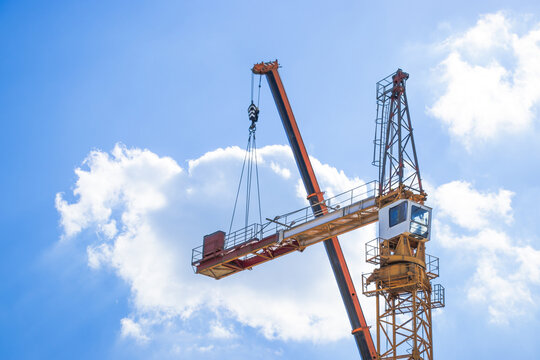 Tower crane with orange and gray structure lifting materials against a bright blue sky with fluffy white clouds in a construction site setting