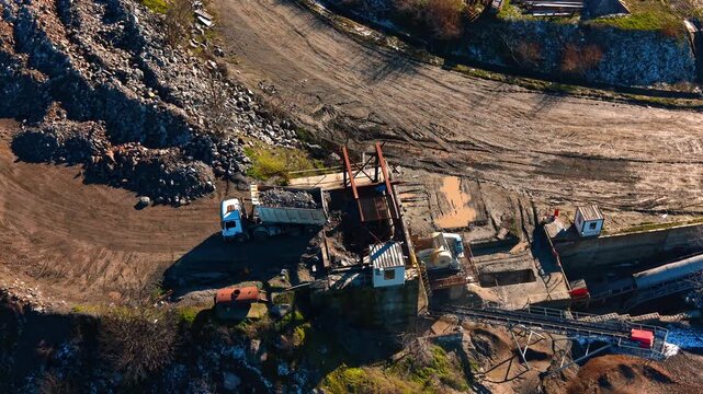 Aerial view of truck unloading stones into industrial rock crusher. Top view of heavy truck dumping rocks at stone processing plant facility during daytime