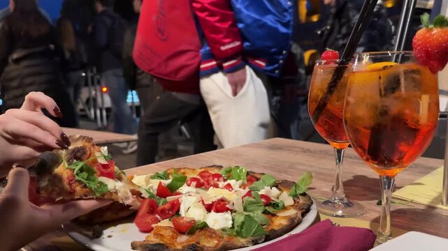 cafe scene near the Colosseum in Rome, Italy. A woman sits at a table by the road, takes a slice of pizza and drinks an aperol with strawberry. Tourists walk past, creating a lively street atmosphere.