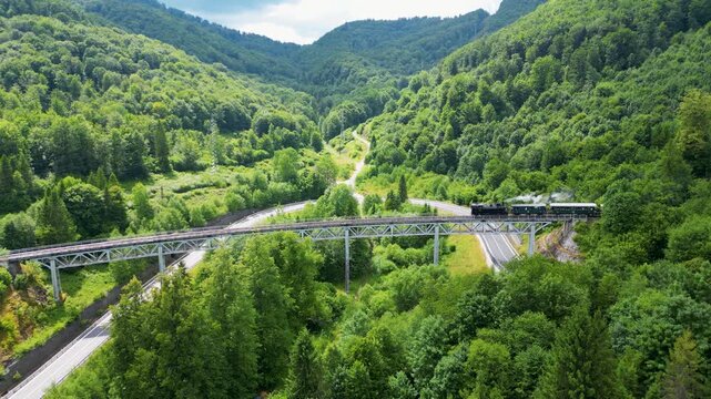 Aerial drone view of historic steam train driving over viaduct and through green fields. Retro steam locomotive puffing smoke in summer nature, Central Europe, 4K, ProRes, 50fps.
