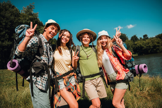 Four friends on a summer backpacking trip by a lake making peace signs and enjoying outdoor adventure and sunshine