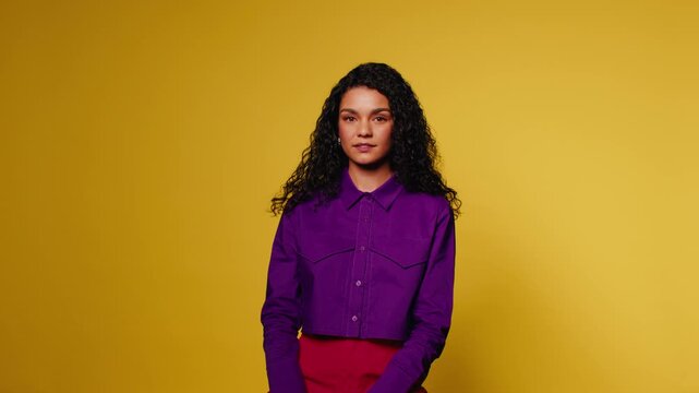 Portrait of confident young woman with long curly hair smiling at the camera against a vibrant yellow background. She displays confidence and elegance.
