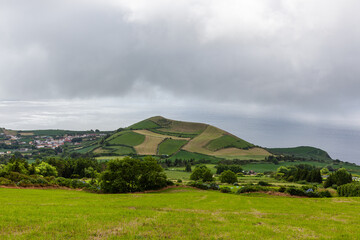 Fototapeta premium Atlantic View, San Miguel Island, Azores, Portugal. Rolling green hills with fields under cloudy sky. Distant view of coastal landscape and ocean. Small town nestled in the valley.