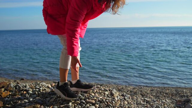 Woman beach jacket standing on a sunny coastline wearing a vibrant pink puffer coat and looking down near the ocean