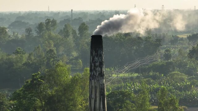 Aerial view of a tall, dark chimney belching grey smoke into the misty sky above vibrant green trees and power lines, Bogura, Rajshahi Division, Bangladesh.