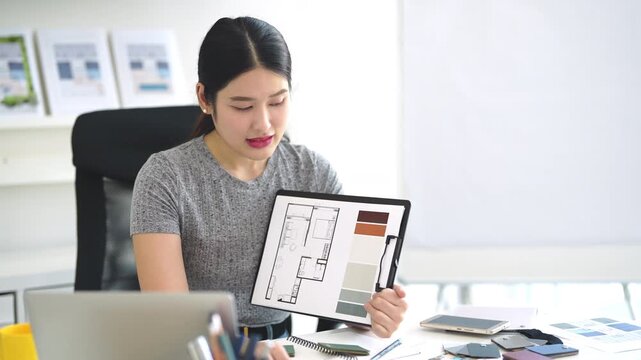Asian Interior designer sitting in front of laptop talking with client on video call, showing architecture floor plan blueprint of residence and pantone. meeting and online conference.