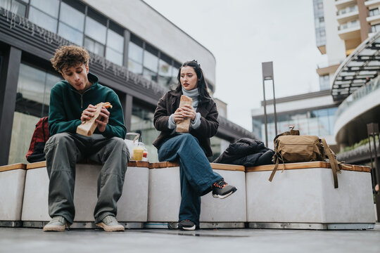 High school friends sit on outdoor benches eating sandwiches and drinking beverages. They relax together in an urban shopping plaza with backpacks at their side.
