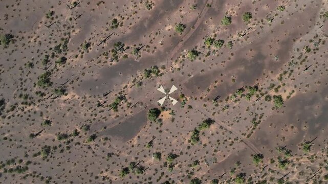 Aerial view of a solitary white windmill contrasting with the arid brown earth and scattered green scrub, featuring subtle dark ground patches, Casa Grande, Arizona, United States.
