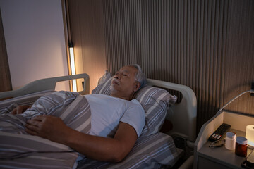 Fototapeta premium Senior man sleeping peacefully in comfortable medical bed during night time feeling calm and relaxed while recovering from illness with warm bedside light