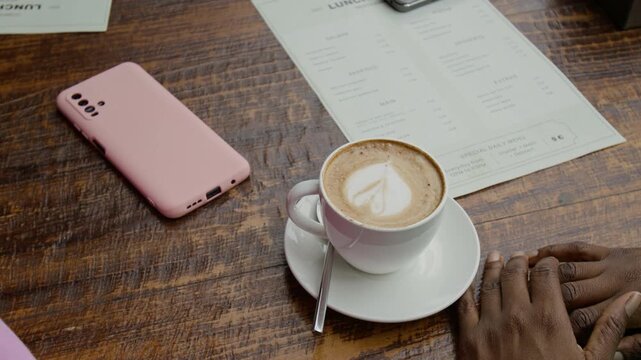 Close-up view of hands of unknown young woman sitting at wooden table featuring smartphone with pink case, menu and white cup of coffee with milk