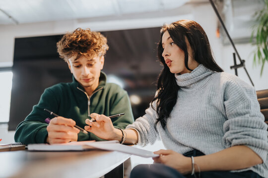 Two students sit at a table working on papers and discussing notes. The pair collaborate on studying and tutoring in a casual classroom setting.