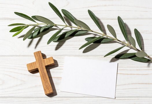 Small wooden cross lying next to an olive branch and a blank white card on a rustic white wooden background, religious concept for Easter, peace, and prayer