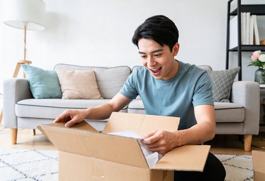 Excited young Asian man opening a cardboard delivery box in a bright living room, feeling surprised and happy with an online shopping purchase and home delivery service.