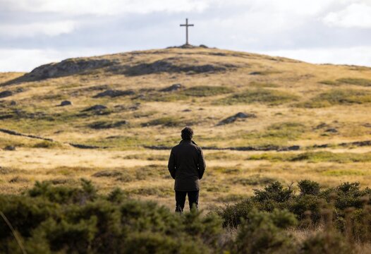 Man standing in a grassy field looking at a large wooden cross on a hill during the day, concept of faith and spirituality for religious contemplation and solitude in nature