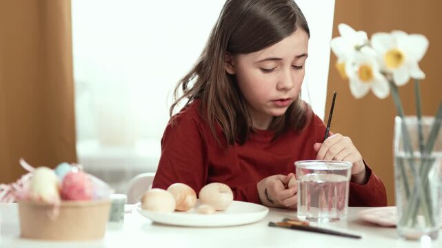 Little girl painting Easter eggs at home, 4K 10-bit cinematic footage of child decorating colorful eggs with watercolor brush for spring holiday, authentic lifestyle.