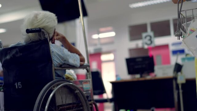 Elderly person sits in a wheelchair inside a hospital. They rest with their hand on their head. Medical equipment and staff are blurred in the background. The scene feels quiet and calm
