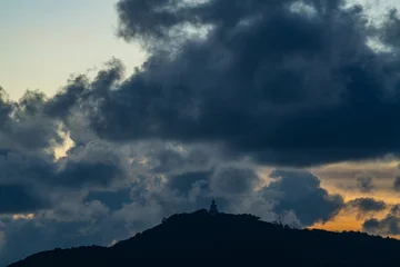Fototapeten Buddha The golden sun sets behind the mountain casting a warm glow around the grand Big Buddha of Phuket. Silhouetted against a radiant sky, the sacred statue radiates peace and spiritual serenity.  © Narong Niemhom