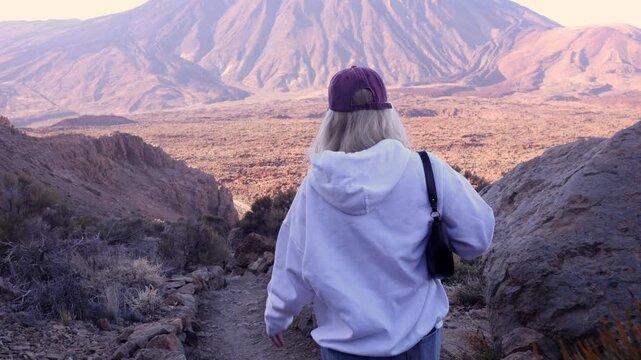 Young blonde woman in white hoodie putting on a cap then setting off along rocky hiking trail with sweeping volcanic valley and mountain view in Tenerife. Authentic adventure travel footage