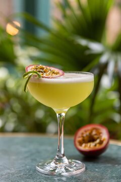 Close up of a tall citronelle yellow green gin cocktail garnished with passion fruit slice and fresh herbs, placed on a textured surface with greenery in the background