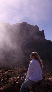 Young blonde woman with red sunglasses sitting on rocky ground, hair blowing in wind, gazing up at dramatic misty volcanic cliff face. Epic adventure travel and lifestyle footage with cinematic light