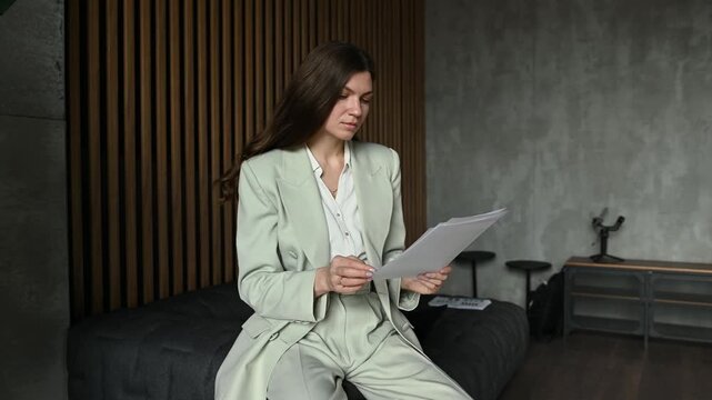 Serious young professional woman sitting in a stylish contemporary office, concentrating on reviewing paperwork, analyzing a contract, or checking a financial report before a business meeting