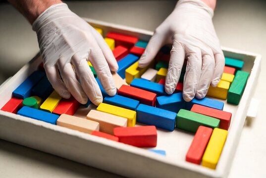 Hands in gloves are arranging colored wooden blocks into a pattern on a tray.