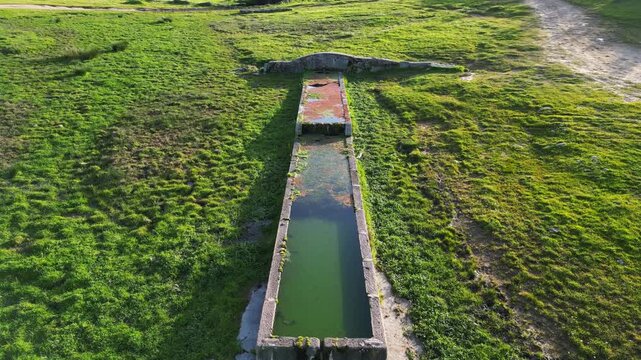 Ascending drone view of a rural livestock water trough revealing the surrounding countryside landscape, green pasture and natural environment.