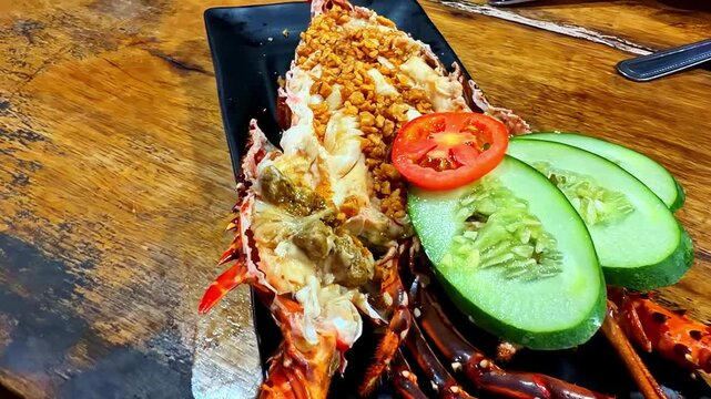 Stuffed lobster presented in its shell with cucumber slices, tomato garnish, and crunchy garlic topping on a black plate over a wooden table.