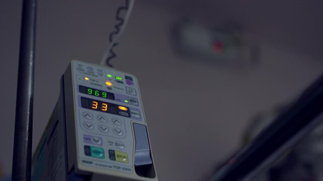 Medical monitor displays vital signs in hospital setting. Digits and lights indicate patient status. Equipment mounted on metal frame. Background shows blurred medical room