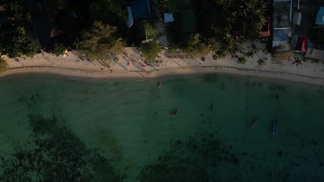 Aerial top-down tracking shot along the beachfront of Koh Lipe showing resort buildings, palm trees, beach loungers, kayaks, and small boats anchored in clear shallow water.