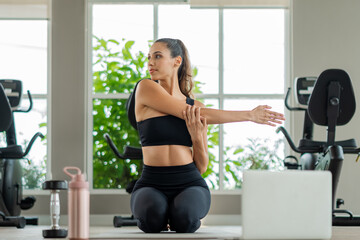 Fototapeta premium Fit woman doing stretching exercises in bright gym. Female athlete doing yoga and fitness exercises with virtual class on computer, digital health and active lifestyle concept.