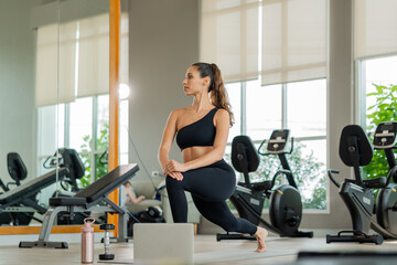 Fototapeta premium Athletic woman performs lunge exercise near mirror in well-lit gym. Female athlete doing yoga and fitness exercises with virtual class on computer, digital health and active lifestyle concept.