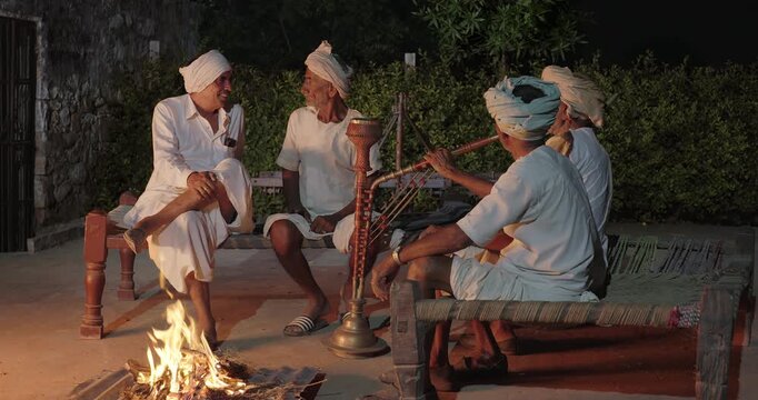 Indian old aged man wear white kurta pajama sitting on woven cot bed enjoy desi life at rural area. Happy four male group talk speak share gossip do hookah hold pipe spend night time village place