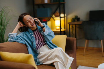 Fototapeta premium Young Korean woman talking on phone while relaxing on sofa at home