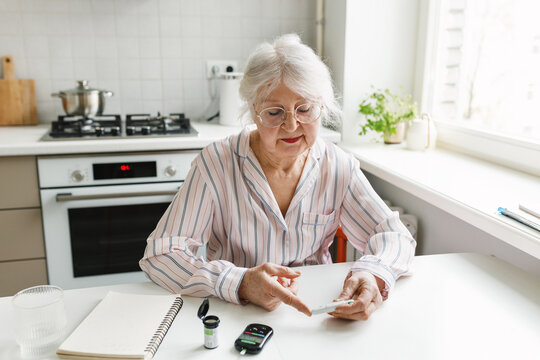 Old lonely Caucasian woman managing diabetes at home performing blood glucose test with lancet and meter checking blood sugar by herself sitting at kitchen table suffering chronic disease