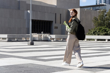 Woman in twenties walking across tiled plaza with green folder, wearing black backpack, copy space © wavebreak3