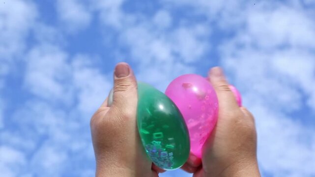 Two hands hold and squeeze transparent green and pink water balloons outdoors against blue cloudy sky in summer, rotating and pressing balloons together showing water movement inside.