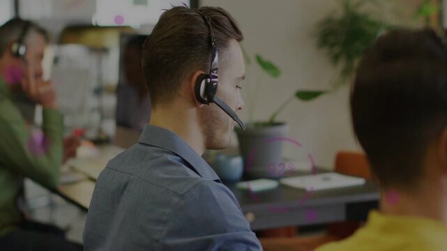 Male call center agent receiving call, speaking into headset and turning and acknowledging coworker