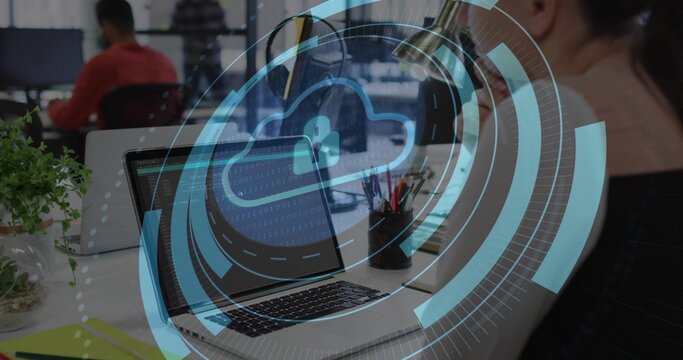 Displaying laptop with cloud lock overlay at open-plan office desk, coworker wearing red shirt