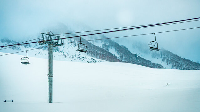 Aerial view of a solitary ski lift and its empty chairs ascending a vast, snow-covered mountain, shrouded in an ethereal mist, Gulmarg, Jammu and Kashmir, India.