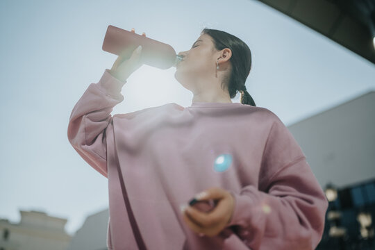 Young woman drinks from a reusable water bottle while standing outdoors. A low-angle view shows her in a pink sweatshirt hydrating in sunlight.