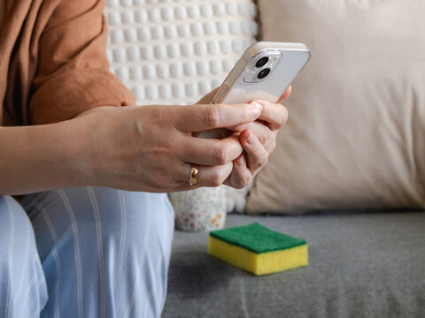 Close-up of wonan sitting on a grey sofa and hands holding smartphone while ordering home cleaning service. Online booking and modern lifestyle concept.