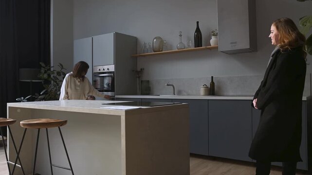 Young mother taking something out of the oven welcomes her daughter returning home, who then leaves as a little girl enters the kitchen. Family life and heartwarming moments in a cozy, stylish home