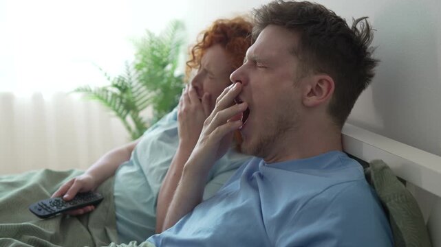 Bored couple watching television and yawning in bed