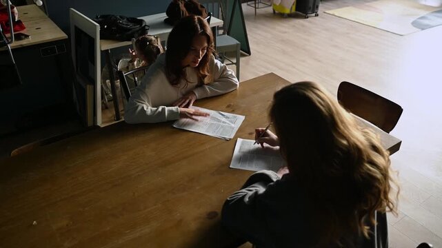 Young woman tutoring a female student, explaining educational material and pointing at a document while the pupil takes notes on a piece of paper, sitting at a wooden table indoors
