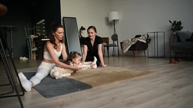 Two skilled female gymnasts teaching and assisting a little girl with stretching exercises, helping her improve flexibility and perform a split on the floor in a modern home studio setting
